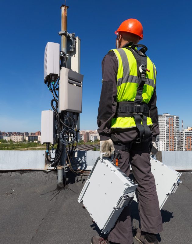 Telecommunication engineer  in helmet and uniform holds telecomunication equipment in his hand and  antennas of GSM  DCS UMTS LTE bands, outdoor radio units, optic fibers, power cables are installed on the roof. Working process of upgrading telecommunication equipment
